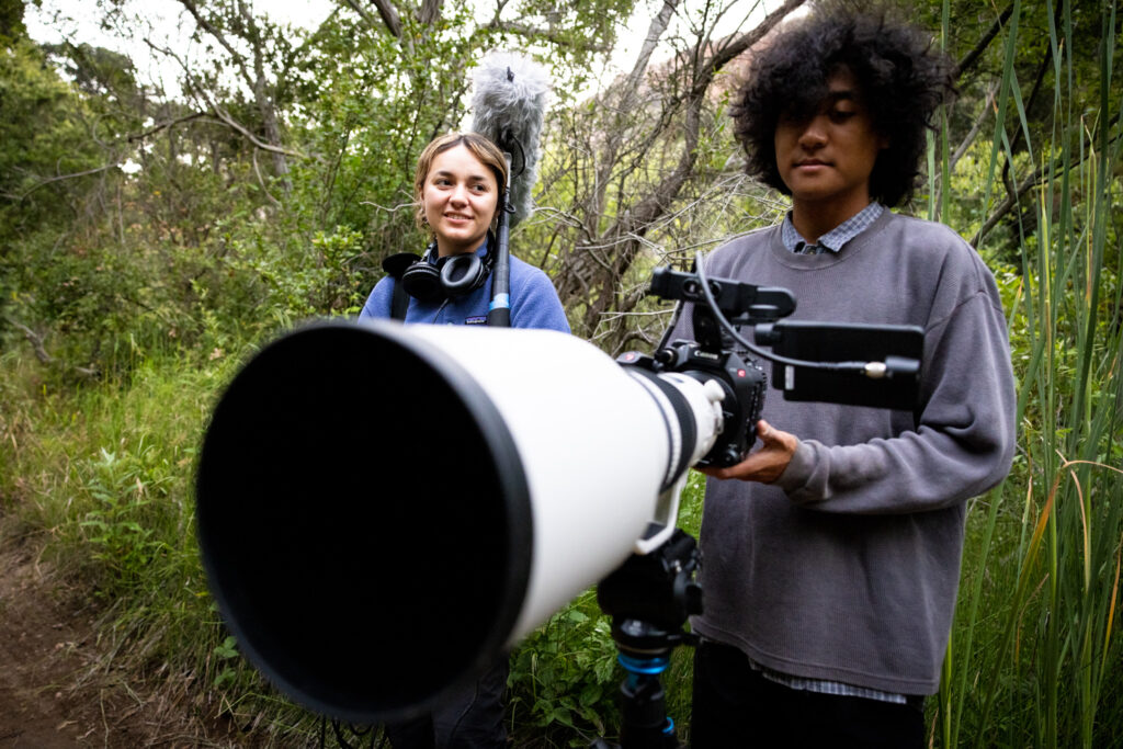 Two students in the jungle, standing behind camera with large zoom in foreground